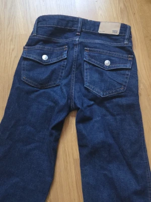 Mörkblå jeans med raka ben - Snygga mörkblå jeans med raka ben och klassisk femficksdesign. Jeansen har silverfärgade knappar på bakfickorna och orangea sömmar som ger en cool kontrast. Tillverkade i denim, perfekta för en avslappnad och trendig look.