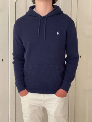 Marinblå hoodie från Polo Ralph Lauren - (Moddelen är 185).        Säljer en marinblå hoodie från Polo Ralph Lauren i ribbad bomullskvalitet. Klassisk huva med dragsko, magficka och broderad logga på bröstet. Perfekt för en chill och stilren look.