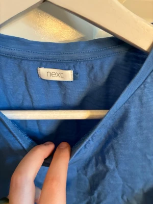 Blå topp med volangärmar från Next - Snygg blå topp från Next med korta volangärmar i broderad spets. Toppen har rund halsringning och är tillverkad i mjuk bomull. Perfekt för dig som vill ha en söt och trendig look med extra detaljer på ärmarna.