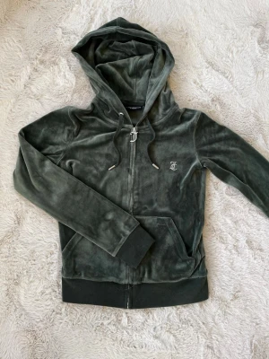 Juicy Couture grön velour hoodie - Mörkgrön velour hoodie från Juicy Couture med dragkedja, huva och fickor. Framsidan har en liten JC-logga och baksidan är dekorerad med glittrande 'Juicy' i stenar. Perfekt för en chill och trendig look.