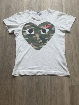 Vit Comme des Garçons PLAY t-shirt - Vit t-shirt från Comme des Garçons PLAY med stort hjärta i camouflage-mönster. Storlek S men sitter som Xs. Skick 8/10, perfekt nu inför sommaren!😀 Hör av dig vid intresse!