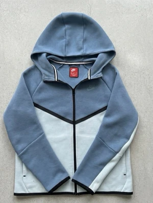 Nike blå och vit hoodie med dragkedja - Nike hoodie i blått och vitt med hel dragkedja framtill och svart detalj längs bröstet. Tröjan har huva och är tillverkad i mjukt material, perfekt för en avslappnad stil. Diskret Nike-logga på bröstet och bak vid nederkanten. 158-170/S