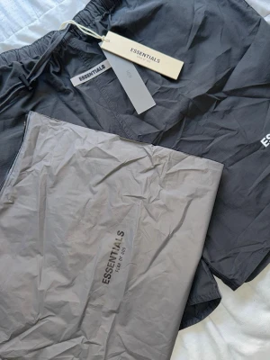 Svarta shorts från Essentials Fear of God - Svarta shorts från Essentials Fear of God med vit logga på benet. De har elastisk midja med snörning och är tillverkade i ett lätt och bekvämt material, perfekta för sommaren. Snygg och enkel design som passar till många olika stilar.