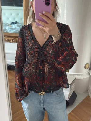 Mönstrad blus med v-ringning - Snygg blus i tunn, transparent chiffong med mörk botten och paisleymönster i rött, orange och grönt. V-ringad med knytning framtill och vida, långa ärmar. Blusen har en lös passform och volang nedtill. Perfekt att styla med jeans för en cool look. Jag är normalt en xs men modellen är L.