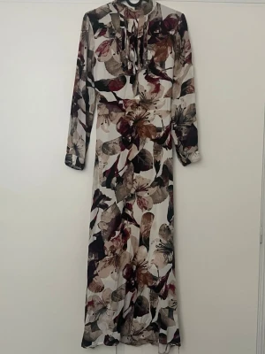 Blommig långklänning med volang - Långklänning med blommigt mönster i beige, vinrött, brunt och svart, blandat färg 🤍🍂❤️🟤Klänningen har långa ärmar, volangdetaljer vid halsen och ett band i midjan. Tillverkad i ett lätt, flytande material som ger en elegant siluett. Obs Det står att den är i storlek L men tror den passar även S 