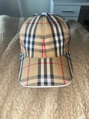 Burberry klassisk rutig keps - Snygg keps från Burberry med det ikoniska beige, svarta, vita och röda rutmönstret. Justerbar rem baktill med Burberry-logga i vitt. Kepsen har böjd skärm och är tillverkad i ett slitstarkt tyg som ger en exklusiv känsla. Pris kan diskuteras 