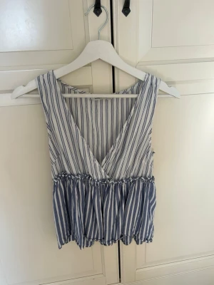 Randig v-ringad topp från ONLY - Säljer en luftig och somrig topp från ONLY i vitt och blått med vertikala ränder. Toppen har v-ringning, är ärmlös och har en volangkant nedtill som ger en avslappnad look. Perfekt för varma dagar och enkel att styla med jeans eller kjol.