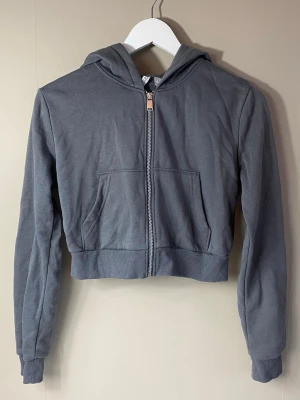 Grå croppad hoodie med dragkedja - Snygg grå croppad hoodie med dragkedja framtill och två fickor. Hoodien har långa ärmar, ribbade muddar och en klassisk huva. Perfekt för en avslappnad och trendig look. 