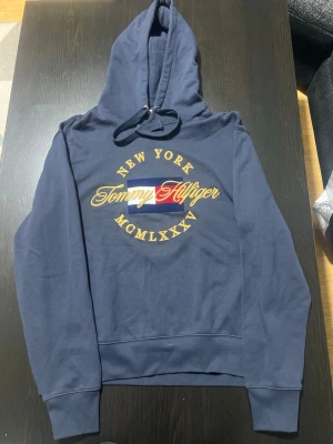 Marinblå hoodie Tommy Hilfiger L - Snygg marinblå hoodie från Tommy Hilfiger med stor huva och dragsko. Framsidan har broderad text 'New York Tommy Hilfiger MCMLXXV' och en flaggdetalj i rött, vitt och blått. Tillverkad i mjukt bomullsmaterial, perfekt för en avslappnad streetwear-look.