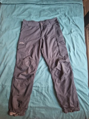 Svarta cargobyxor från Lundhags - Snygga frilufts/vandringsbyxor från Lundhags med flera praktiska fickor på sidorna och bak. Byxorna har resår vid bensluten och förstärkta sömmar över knäna. Slitstarkt material och sista bilden visar vad byxorna ligger på i nypris. Säljes i befintligt skick.