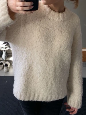 Krämvit fluffig stickad tröja - Mysig krämvit stickad tröja med fluffig struktur och rund hals. Tröjan har en avslappnad passform och långa ärmar, perfekt för kalla dagar. Passar till både jeans och kjol för en chill look.