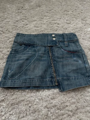 Blå jeanskjol med broderi - Säljer en kort blå jeanskjol med snyggt vitt broderi på både fram- och baksidan. Kjolen har två knappar i midjan, bälteshällor och dragkedja framtill. Klassisk femficksmodell med coola detaljer och en liten slits framtill.