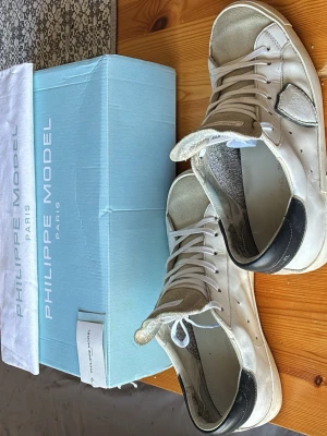 Vita Philippe Model sneakers - Snygga vita sneakers från Philippe Model med svarta detaljer på hälen och beige mocka på tån och hälen. Klassisk låg modell med vita skosnören och diskret logga på baksidan. Perfekta för dig som gillar stilrena och trendiga skor.