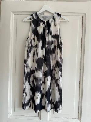 Svartvit ärmlös klänning H&M - Säljer en ärmlös klänning från H&M med svartvitt abstrakt mönster. Klänningen har rund halsringning med liten knappdetalj framtill och är i ett mjukt, lätt material som faller snyggt. Perfekt för dig som gillar en stilren men unik look.