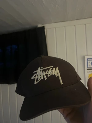 Brun Stüssy keps med broderad logga - Säljer en svart keps från Stüssy med deras ikoniska vita broderade logga framtill. Kepsen har en böjd skärm med grön undersida och justerbar snapback baktill. Perfekt för dig som gillar streetwear och vill ha en stilren accessoar.