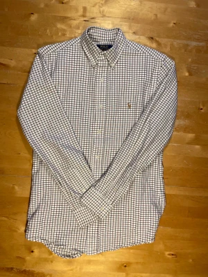 Rutig skjorta från Polo Ralph Lauren - Klassisk vit skjorta med blå rutor från Polo Ralph Lauren. Skjortan har lång ärm, knappar framtill och en liten broderad logga på bröstet. Tillverkad i bomull och har en tidlös, normal passform som funkar till många olika stilar.