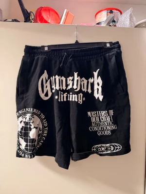 Svarta Gymshark shorts med tryck - Säljer ett par svarta shorts från Gymshark med stora vita tryck framtill, bland annat logga, text och grafiska detaljer. Shortsen har resårmidja med dragsko och sidofickor. Perfekta för gymmet eller chill. Materialet känns mjukt och bekvämt.