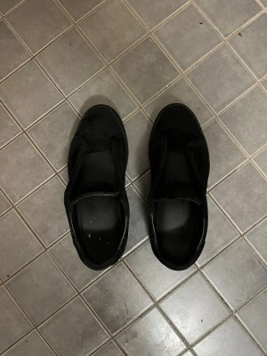  Axel Arigato laceless  - Stilrena svarta sneakers från Axel Arigato i mocka med diskret logga på sidan. Klassisk låg modell med svart sula och minimalistisk design som passar till det mesta. Skorna har en clean look utan snören för en modern vibe.