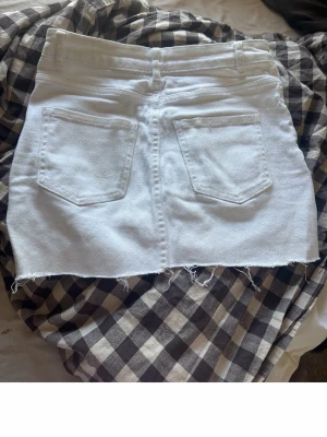 Vit jeanskjol med rå kant - Säljer en vit kort jeanskjol med rå, fransig kant nertill och två bakfickor. Kjolen har klassisk femficksdesign och är perfekt för en avslappnad och trendig look.