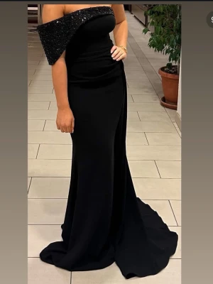 Elegant svart långklänning - Säljer en elegant svart långklänning med figurnära passform och släp. Klänningen har en klassisk och stilren design utan synliga detaljer eller mönster. Perfekt för dig som vill ha en tidlös look till speciella tillfällen.