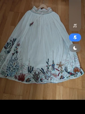 Vit långklänning med broderi - Unik vit långklänning med färgglada broderade undervattensmotiv längs fållen. Klänningen har snörning vid halsen och en lös, flowy passform. Perfekt för dig som gillar detaljer och bohemisk stil.