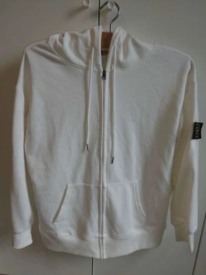 Vit zip hoodie från Stone Island - Säljer en clean vit hoodie från Stone Island med dragkedja framtill och klassisk huva med dragsko. Hoodien har känguruficka och den ikoniska svarta patchen med gult kompassmärke på ärmen. Tillverkad i mjuk bomull för skön vardagsstil. 