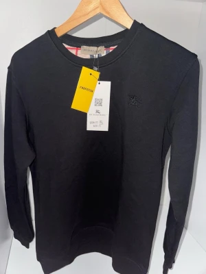 Svart tröja  från Burberry - Snygg svart tröja  från Burberry med klassiskt rutigt mönster på insidan av kragen och diskret broderad logga på bröstet. Tröjan  har rund hals och korta ärmar, perfekt för en clean och stilren look. De på mitten av tröjan är lins från kameran. Finns 2 kvar storlek S och M inne.