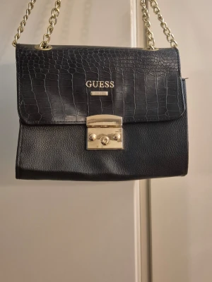 Svart handväska från Guess med kedja - Snygg svart handväska från Guess med guldfärgad kedjerem och krokopräglat lock. Väskan har ett elegant metallspänne framtill och Guess-logga i guld. Materialet är konstläder med struktur och slät botten. Perfekt för dig som gillar trendiga accessoarer.