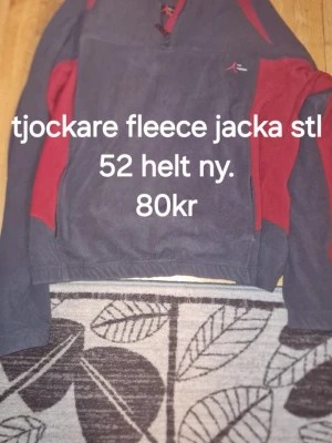 Mörkblå och röd fleecejacka - Mysig och tjock fleecejacka i mörkblått med röda detaljer. Jackan har lång ärm, halv dragkedja framtill och en liten logga på bröstet. Perfekt för kyliga dagar och enkel att dra på sig.