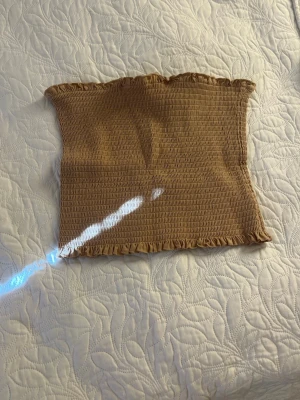 Beige smockad tubtopp med volang - Trendig beige tubtopp med smockad struktur och volangkant upptill och nedtill. Perfekt att styla till jeans eller kjol för en enkel men snygg look. Mjuk och stretchig känsla.