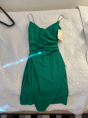 Grön omlottklänning med smala band - Snygg grön omlottklänning med smala axelband och v-ringning. Klänningen har en figurnära passform och är tillverkad i ett mjukt, lätt material som passar perfekt till varmare dagar. Stilren och enkel design som ger en elegant look.