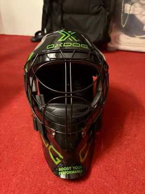 Oxdog innebandymålvaktshjälm svart/grön - Snygg innebandymålvaktshjälm från Oxdog i svart med gröna detaljer och text. Hjälmen har ett robust galler framtill, justerbara remmar och vadderad insida för extra komfort. Perfekt för dig som vill ha både skydd och stil på planen.