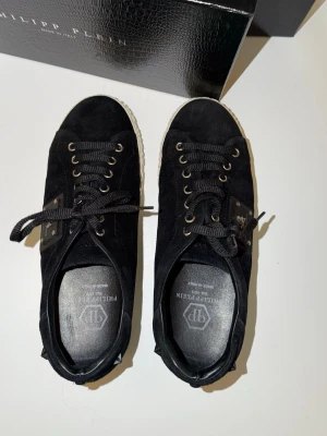 Svarta Philipp Plein sneakers i mocka - Svarta låga sneakers från Philipp Plein i mocka med svarta skosnören och guldfärgade metalldetaljer. Vit sula med tydlig Philipp Plein-logga på sidan. Snygg och stilren design med exklusiv känsla.