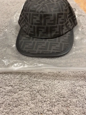 Svart keps - Snygg svart keps med det klassiska FF-monogrammet över hela kepsen. Kepsen har böjd skärm och är tillverkad i ett slitstarkt tyg med läderdetalj på skärmen. Perfekt accessoar för dig som gillar exklusiv streetwear.