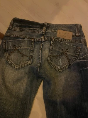Blå bootcut jeans från Eight - Säljer ett par blå bootcut jeans från Eight med slitningar och snygga broderade detaljer på bakfickorna. Jeansen har låg midja och klassisk femficksmodell. Materialet är denim i bomull med en skön passform.