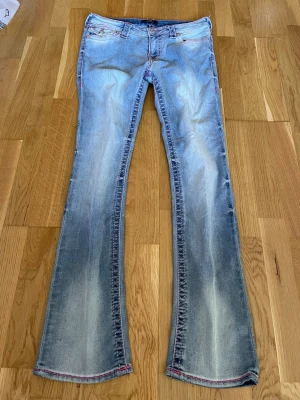 Bootcut ljusblå jeans med röda sömmar - Säljer ett par ljusblå bootcut jeans med snygga slitningar och röda kontrastsömmar. Jeansen har klassisk femficksmodell och är tillverkade i denim med lite stretch. Perfekta för en avslappnad och trendig look.