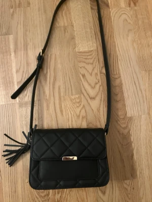 Svart axelväska från Anna field  - Snygg svart axelväska med quiltat mönster och justerbar axelrem. Väskan har en dekorativ tofs på sidan och ett metallspänne framtill. Perfekt storlek för det viktigaste och ger en trendig touch till din outfit. Pris kan diskuteras!