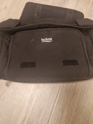 Svart barnvagn väskan från Britax - Säljer en svart datorväska från Britax med justerbar axelrem och flera fack med dragkedja. Väskan är tillverkad i slitstarkt tygmaterial och har en enkel, stilren design med Britax-logga på framsidan. Perfekt för att bära laptop och tillbehör.