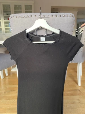 Svart ribbad klänning från Lindex - En svart ribbad kortklänning från Lindex med korta ärmar och söta volangkanter. Klänningen har en enkel och stilren design som passar perfekt till många olika tillfällen. Köparen står för frakt.