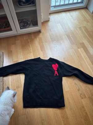 Svart stickad tröja från Ami Paris - Säljer en svart stickad tröja från Ami Paris med röd hjärt- och A-detalj på bröstet. Tröjan har rund halsringning, långa ärmar och ribbade muddar. Perfekt för dig som gillar stilrena plagg med en twist.