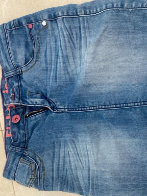 Blå jeans med färgglada detaljer - Säljer ett par blå jeans med slitningar och färgade nitar i rosa och gult. Jeansen har klassisk femficksmodell, dragkedja och synliga sömmar. Insidan av linningen har ett rosa tryck. Perfekta för dig som gillar unika detaljer!