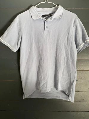 Ljusblå pikétröja från 157 - Klassisk ljusblå pikétröja från 157 med krage och knappar framtill. Tillverkad i mjuk bomull och har korta ärmar. Perfekt för en avslappnad och stilren look.