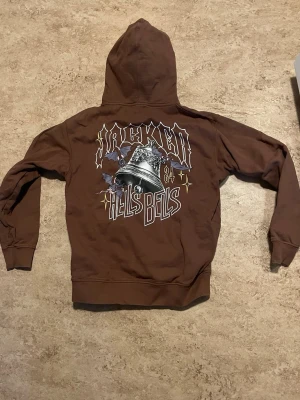 Jacker Hoddie “Hell’s Bells” - En ljusbrun hoddie i märket Jacker storlek M. Trycket ”Hell’s Bells” finns på baksidan samt trycket ”Jacker” på framsidan.