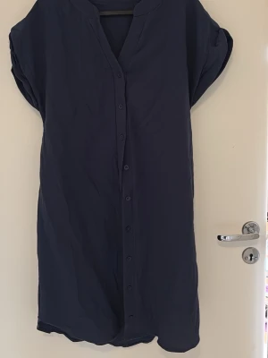 Ny! Mörkblå skjortklänning med knappar - En enkel och stilren mörkblå skjortklänning med knappar längs hela framsidan. Klänningen har korta ärmar och en avslappnad passform, perfekt för en chill och bekväm look. Materialet känns lätt och svalt, vilket gör den skön att bära under varmare dagar.