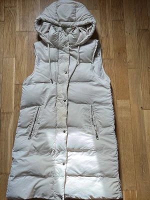 Lång beige dunväst med huva - Snygg lång dunväst i beige med stor huva och dragsko. Västen har två dragkedjeförsedda fickor framtill och stängs med tryckknappar. Perfekt för lager på lager och ger en trendig look till höst och vinter.