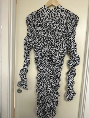 Långärmad leopardmönstrad klänning - Säljer en långärmad, figurnära klänning med svartvitt leopardmönster. Klänningen har hög hals, rynkade detaljer längs hela plagget och puffiga ärmar. Perfekt för dig som vill sticka ut med en trendig och unik look.