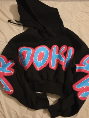 Svart hoodie med färgglad text - Säljer en svart hoodie med stor huva och färgglad graffiti-inspirerad text i blått och rosa på bröstet och ärmarna. Hoodien har en croppad passform och ribbade muddar. Perfekt för dig som gillar streetwear och vill sticka ut.