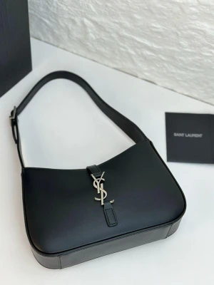 Svart axelremsväska från Saint Laurent - Snygg svart handväska i slätt läder från Saint Laurent med ikoniskt YSL-spänne i silver framtill. Väskan har en stilren, rektangulär form och justerbar axelrem. Perfekt för dig som gillar klassisk och lyxig design.