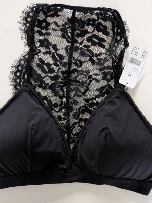 Cubus Svart spetsbralette Behå Storlek M - Snygg svart bralette från H&M med triangelformade kupor och bred spetsdetalj i ryggen. Axelbanden är justerbara och materialet är en mix av mjuk stretch och transparent spets med blommönster. Perfekt för dig som gillar en trendig och bekväm look.