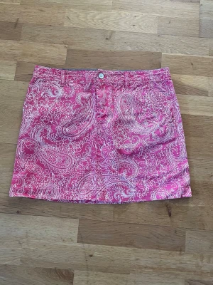 Rosa kortkjol - Säljer en rosa kortkjol med vitt paisleyinspirerat mönster. Kjolen har klassisk femficksdesign, bälteshällor och stängs med knapp och dragkedja framtill. Perfekt för dig som vill sticka ut med färg och mönster i garderoben.